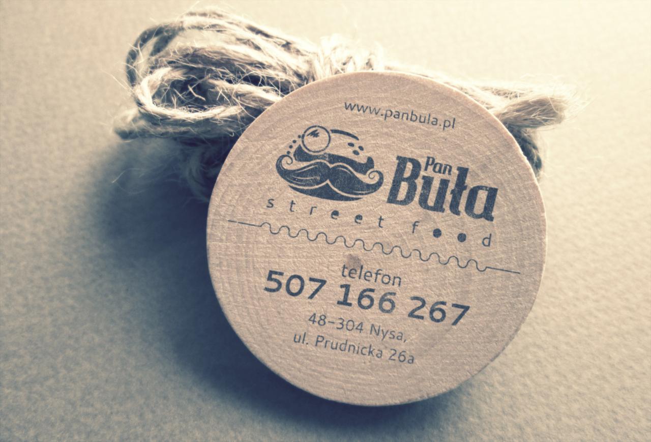 Okrągła, drewniana wizytówka firmy Pan Buła street food z adresem strony www, numerem telefonu i adresem, obok leży sznurek jutowy.