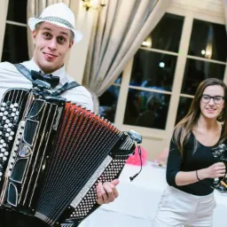 Młody mężczyzna w białym kapeluszu i muszce gra na akordeonie marki Crucianelli, pokazując język, obok kobieta z tamburynem, w tle okna i goście weselni.