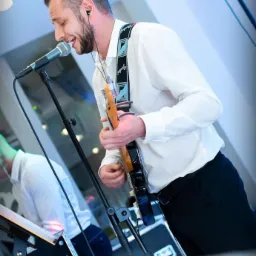 Wokalista z gitarą elektryczną śpiewa do mikrofonu podczas występu na scenie, w tle widoczny drugi muzyk.