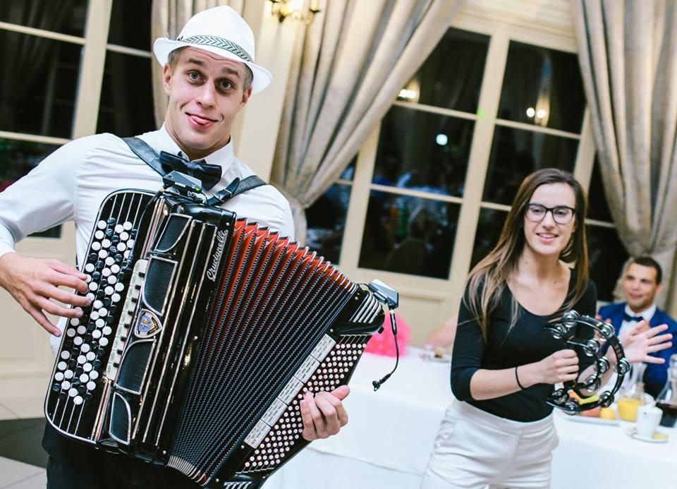 Młody mężczyzna w białym kapeluszu i muszce gra na akordeonie marki Crucianelli, pokazując język, obok kobieta z tamburynem, w tle okna i goście weselni.