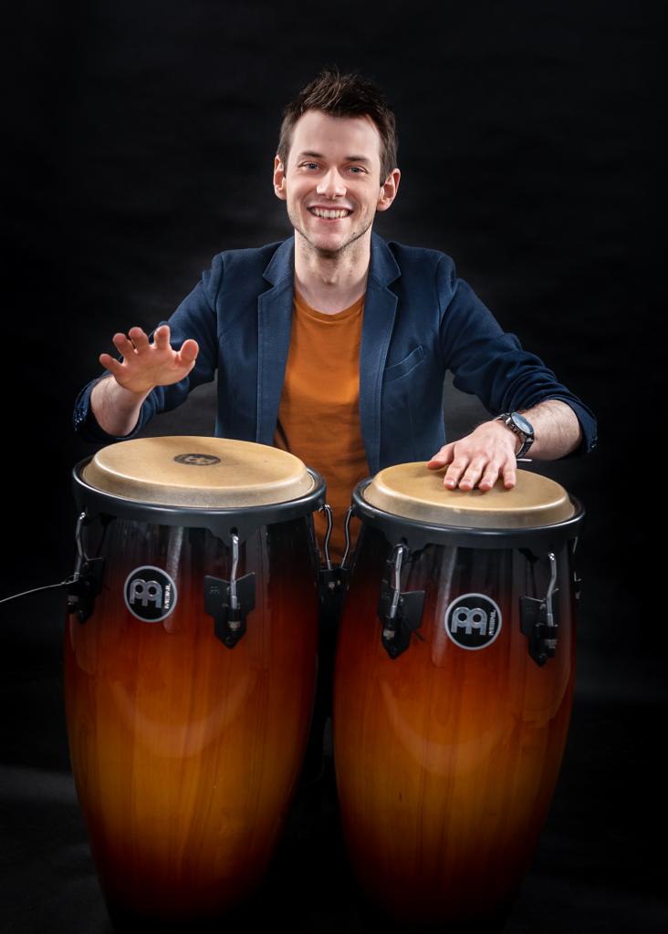 Uśmiechnięty muzyk w niebieskiej marynarce grający na dwóch bębnach conga marki Meinl na czarnym tle.
