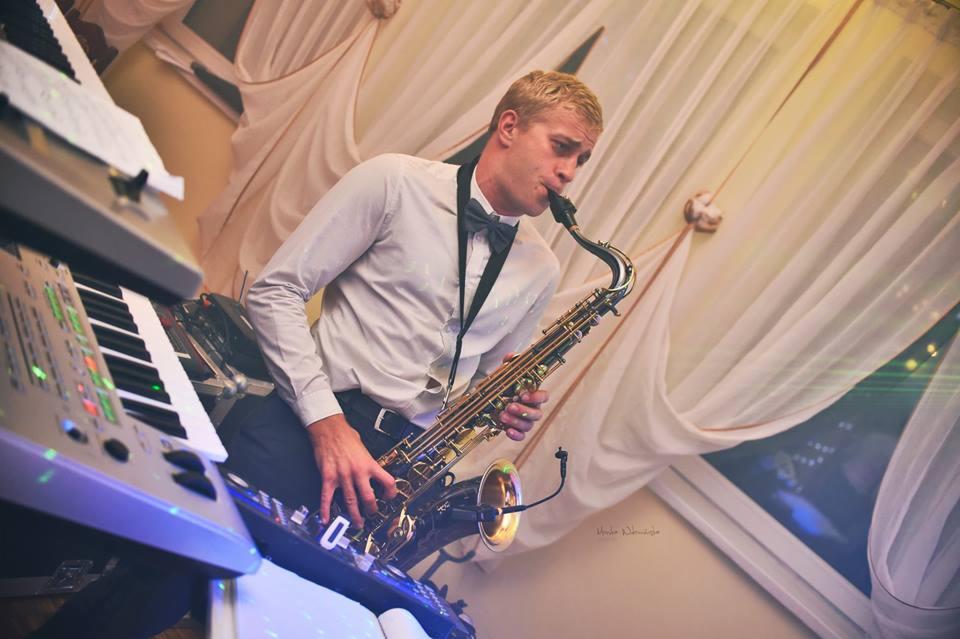 Saksofonista w muszce grający na saksofonie tenorowym na tle białych zasłon i klawiszy syntezatora.