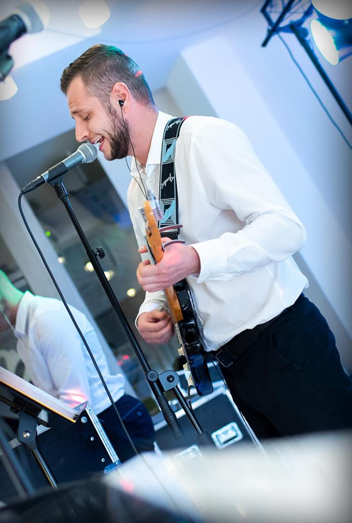 Wokalista z gitarą elektryczną śpiewa do mikrofonu podczas występu na scenie, w tle widoczny drugi muzyk.