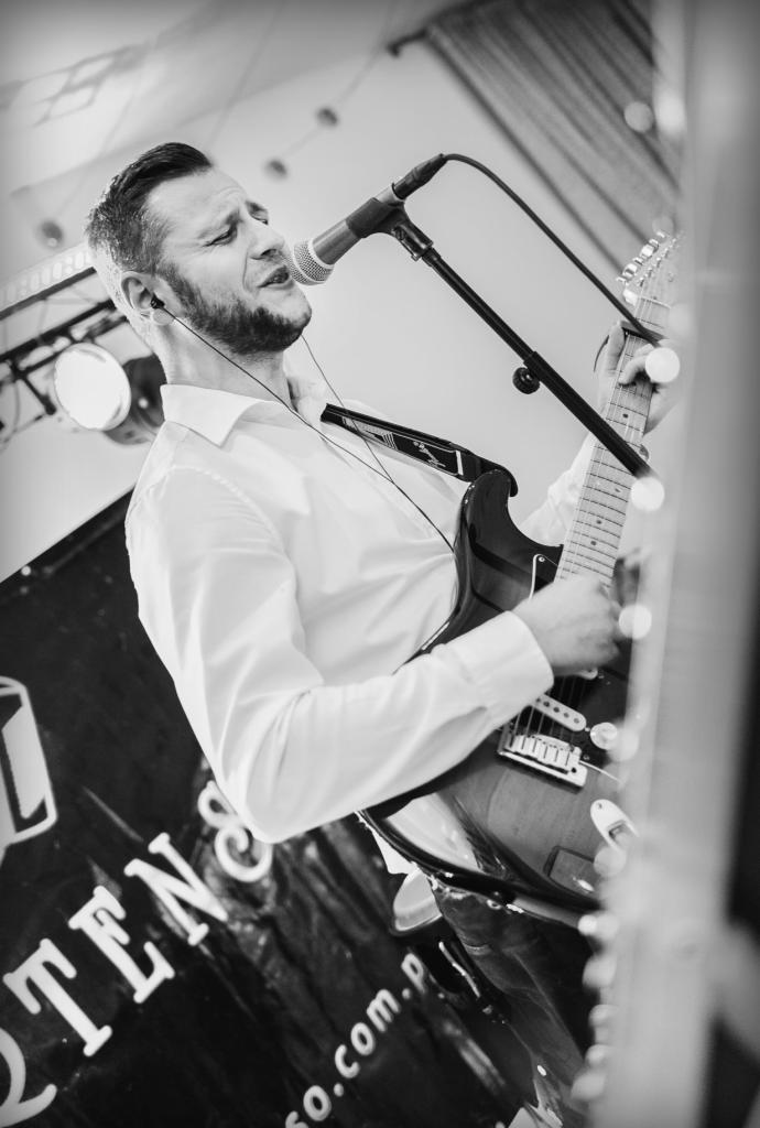 Czarno-białe zdjęcie muzyka w białej koszuli, grającego na gitarze elektrycznej i śpiewającego do mikrofonu, z widocznymi słuchawkami w uszach, na tle sceny z oświetleniem i fragmentem baneru...