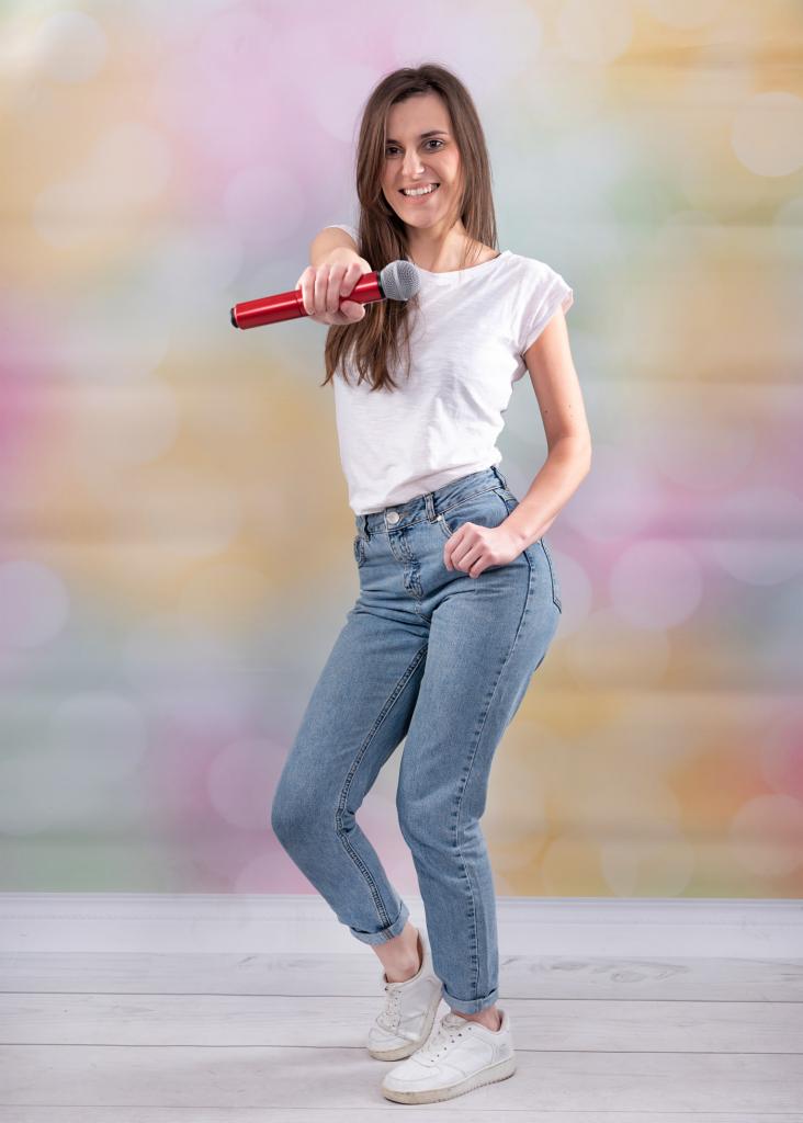 Uśmiechnięta kobieta w jeansach i białej koszulce trzymająca czerwony mikrofon na tle kolorowego, rozmytym bokeh.