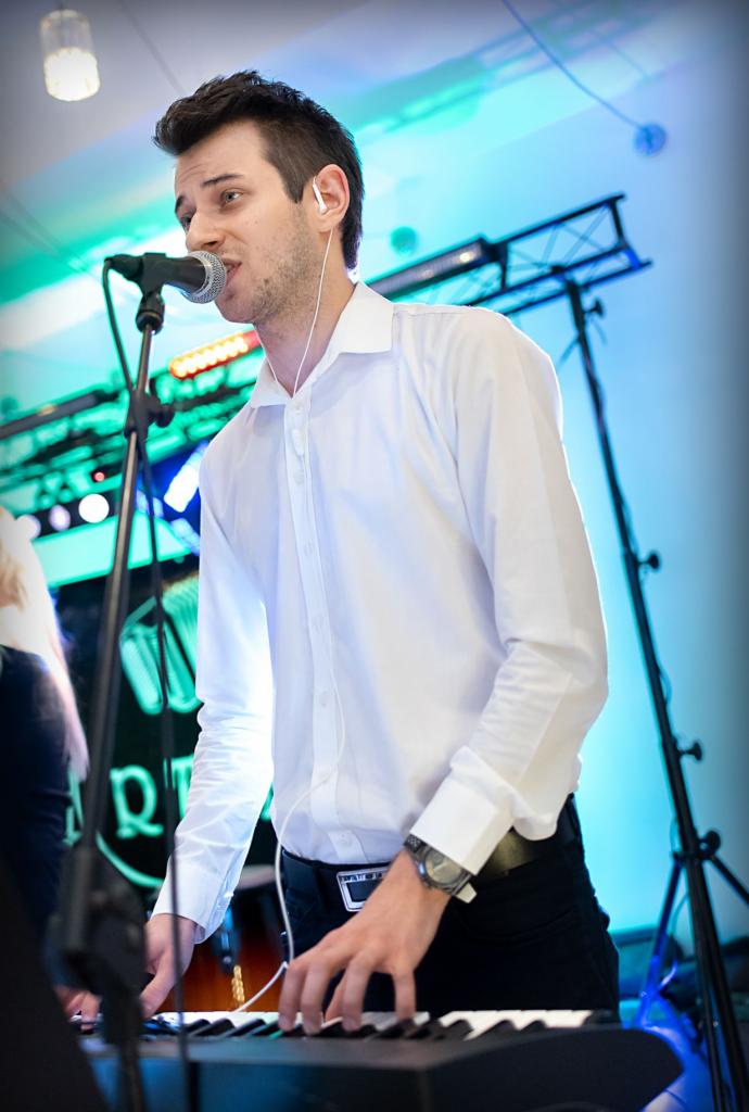 Pianista w białej koszuli, z mikrofonem i słuchawkami, grający na keyboardzie podczas występu na tle oświetlenia scenicznego.