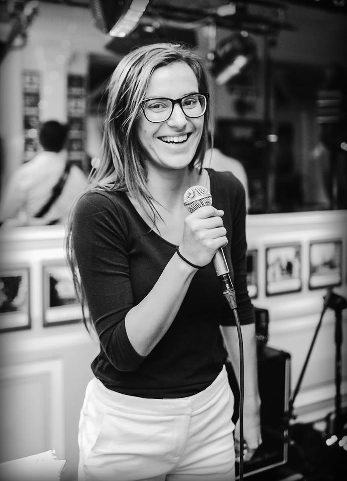 Uśmiechnięta kobieta w okularach trzyma mikrofon na tle sali z dekoracjami ściennymi.