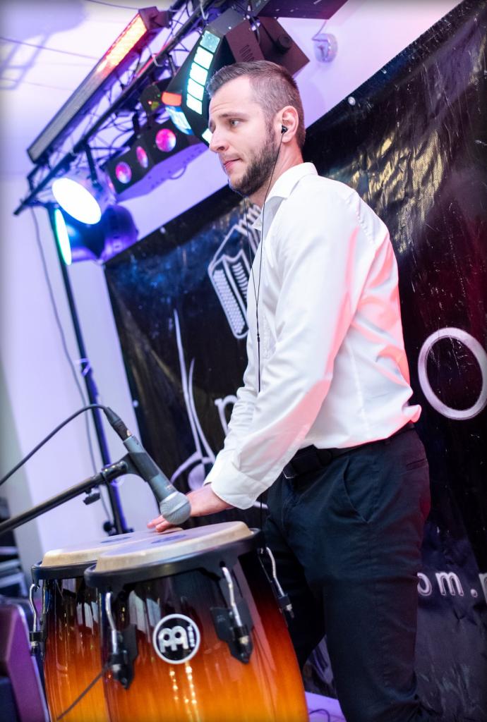 Perkusjonista grający na bębnach conga na scenie, oświetlenie sceniczne w tle, mikrofon przed bębnami.