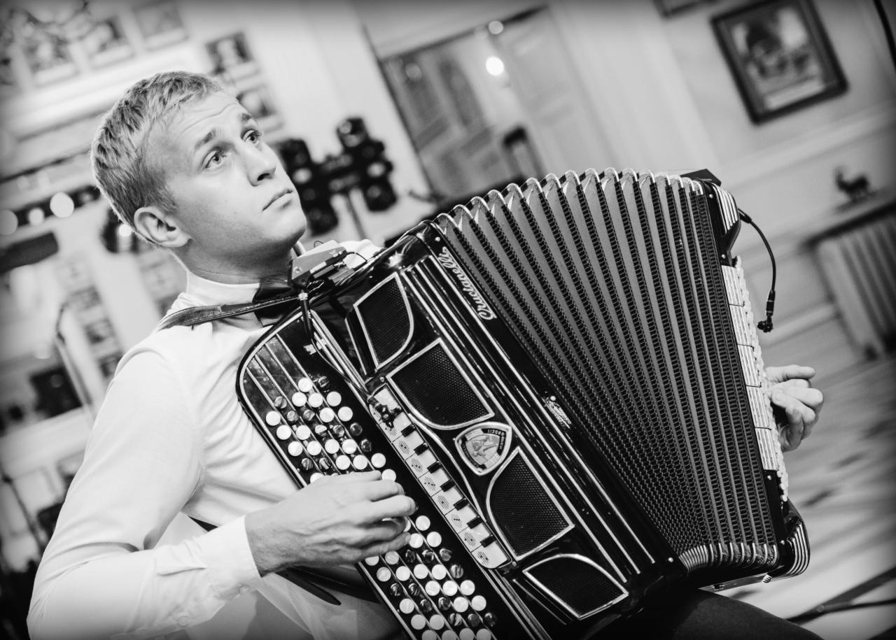Czarno-białe zdjęcie młodego mężczyzny w białej koszuli i muszce, grającego na akordeonie marki Crucianelli, z widocznym mikrofonem przypiętym do instrumentu, w tle rozmyte wnętrze z obrazami...