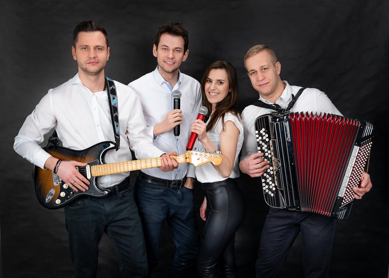 Portret studyjny czteroosobowego zespołu: gitarzysta z gitarą typu sunburst, dwóch wokalistów z mikrofonami i akordeonista z czerwono-czarnym akordeonem.