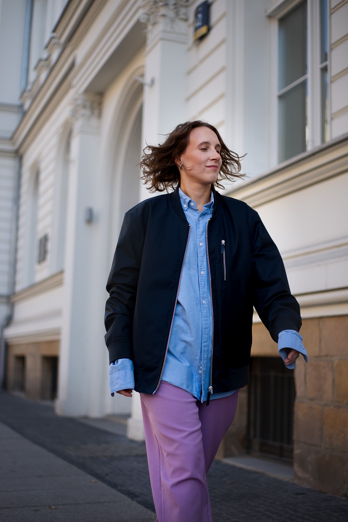 Kobieta w bomberce, błękitnej koszuli i liliowych spodniach idzie ulicą. W tle biały budynek z kolumnami. Modna stylizacja na co dzień, swobodny look.