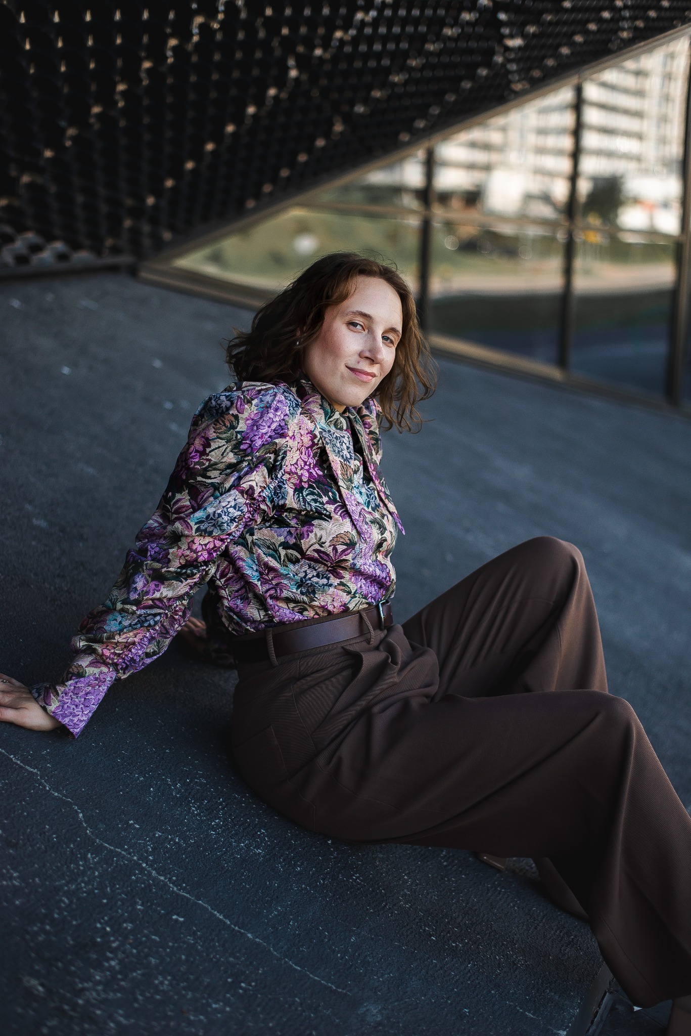 Kobieta siedzi w brązowych spodniach i kwiecistej bluzce pod nowoczesnym budynkiem. Uśmiechnięta, patrzy w obiektyw. Stylizacja z charakterem, naturalny makijaż.