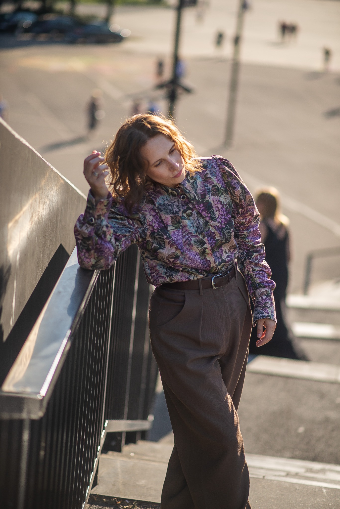Kobieta w kwiecistej bluzce i szerokich spodniach pozuje oparta o barierkę schodów w słoneczny dzień. Stylizacja codzienna z nutą elegancji.