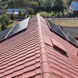 Panele słoneczne zamontowane na skośnym dachu z czerwonej dachówki ceramicznej, widok z góry na tle zabudowań i zieleni.