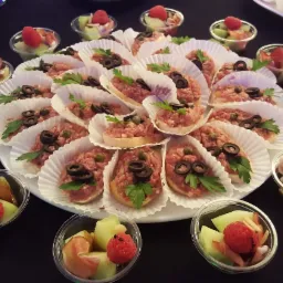 Elegancko ułożone tartinki z tatarem wołowym, oliwkami i kaparami na białych papilotkach, otoczone małymi miseczkami z melonem, szynką parmeńską i malinami, całość na czarnym tle.