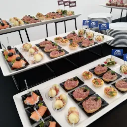 Elegancko podane przekąski cateringowe na białych paterach z flagami firmowymi, widoczne tartaletki, mini-kanapeczki z tatarem, roladki z grilla i łososia, oraz stosy białych talerzy.