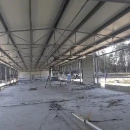 Wnętrze budowanej hali z konstrukcją stalową dachu, widoczne okna i brama segmentowa, betonowa posadzka z resztkami materiałów budowlanych.