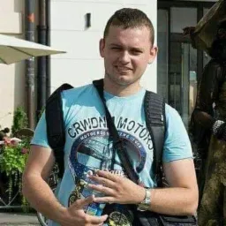 Uśmiechnięty mężczyzna w niebieskiej koszulce z motywem motocyklowym, stojący na zewnątrz z torbą na ramię i plecakiem, prawdopodobnie programista podczas przerwy.