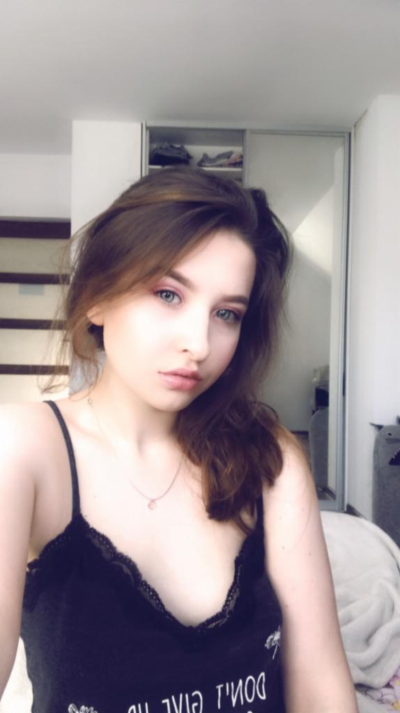 Portret młodej kobiety z delikatnym makijażem w pokoju z białą szafą w tle.