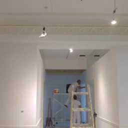 Dwóch malarzy na rusztowaniu i drabinie odnawia wnętrze z ozdobnym gzymsem sufitowym, zabezpieczone folią okno po prawej stronie.