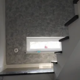 Ściana w korytarzu wykończona dekoracyjnym tynkiem w odcieniach szarości, widoczne schody z czarnymi stopniami i białą balustradą, małe okno z parapetem.