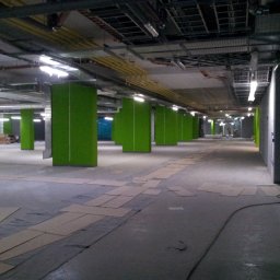 MAVI Piotr Dźwięga - Wnętrze biurowe w trakcie prac wykończeniowych; widoczne zielone kolumny, odsłonięty sufit z instalacjami, podłoga zabezpieczona kartonem i folią.