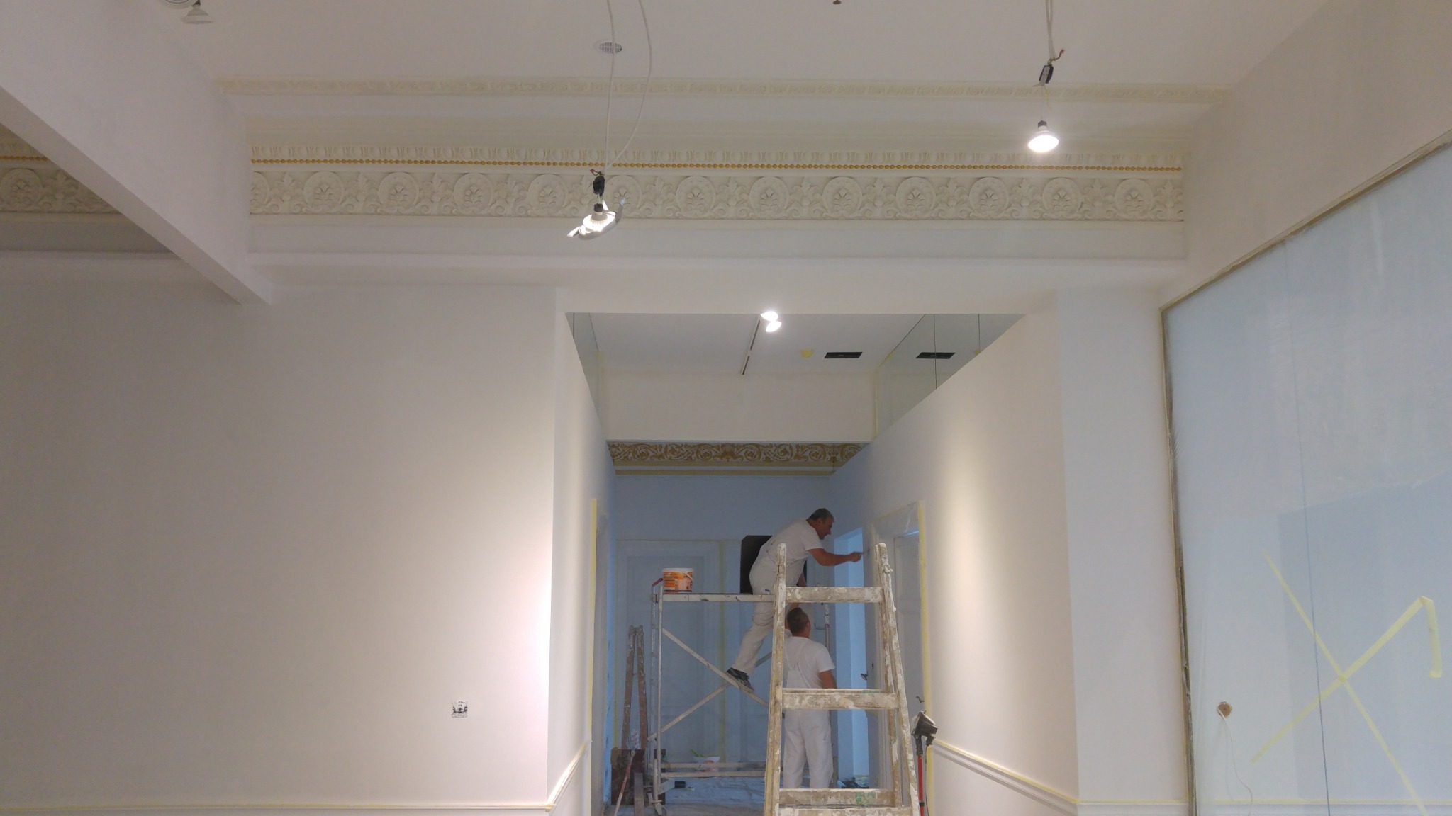Dwóch malarzy na rusztowaniu i drabinie odnawia wnętrze z ozdobnym gzymsem sufitowym, zabezpieczone folią okno po prawej stronie.