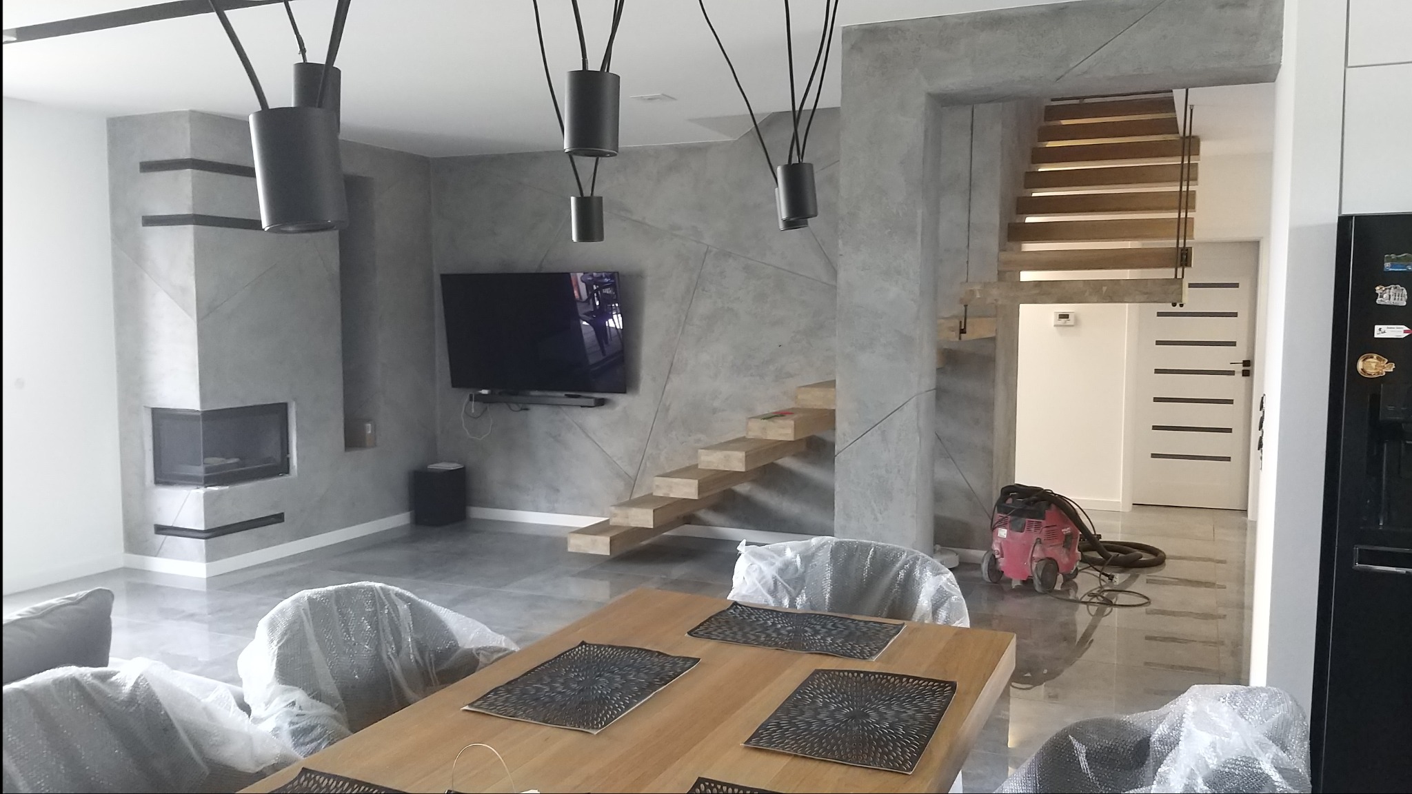 Nowoczesne wnętrze po remoncie: szara ściana z geometrycznym wzorem, drewniane schody bez poręczy, kominek, telewizor, stół z krzesłami zabezpieczonymi folią oraz odkurzacz na podłodze.