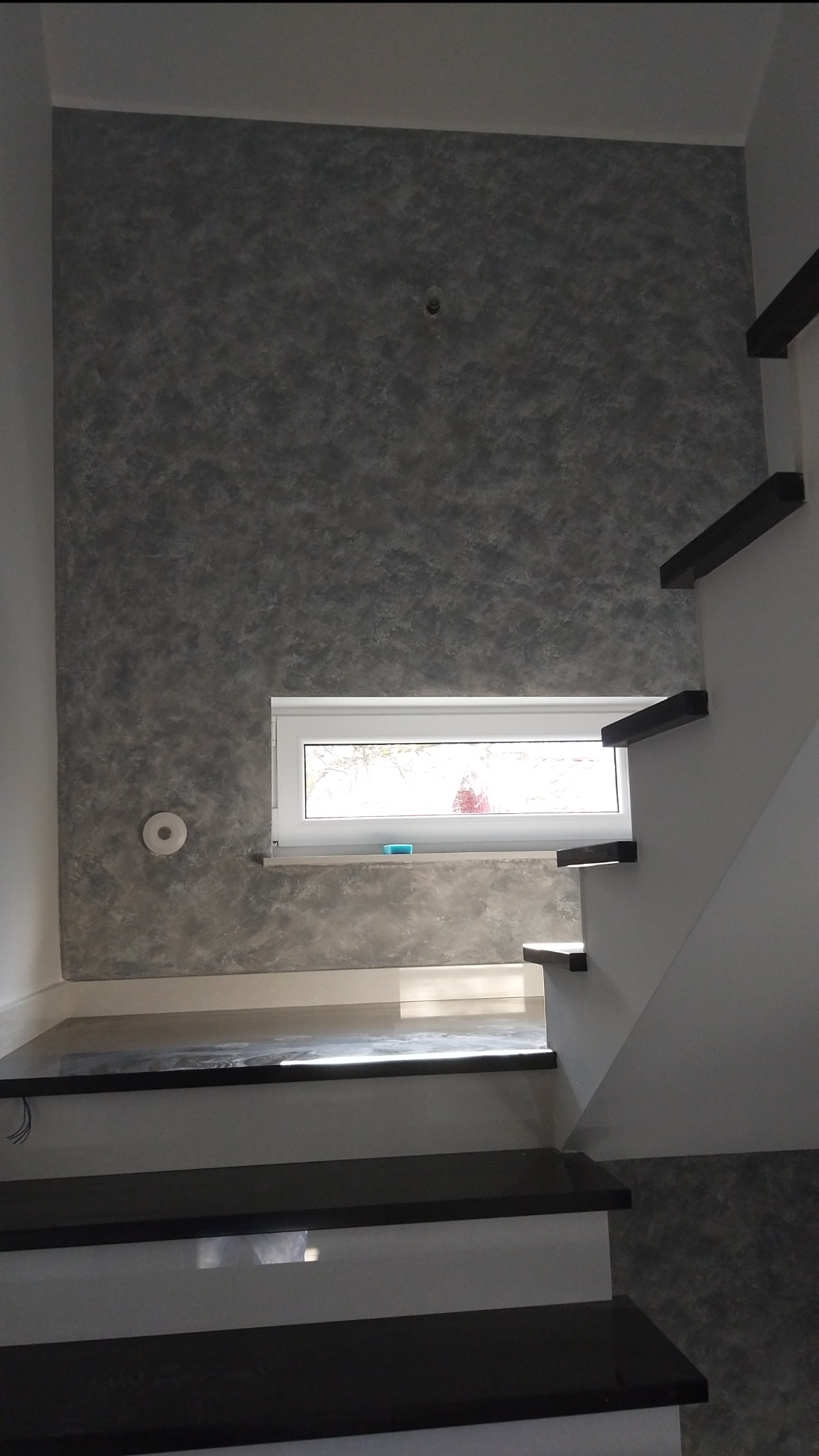 Ściana w korytarzu wykończona dekoracyjnym tynkiem w odcieniach szarości, widoczne schody z czarnymi stopniami i białą balustradą, małe okno z parapetem.