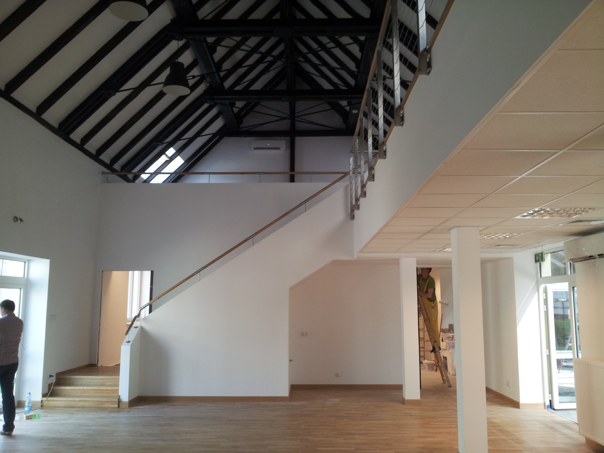 Przestronne wnętrze w trakcie wykańczania: białe ściany, drewniana podłoga, schody z metalową balustradą, widoczny pracownik na drabinie, surowy strop z drewnianymi belkami, światło wpadające przez...