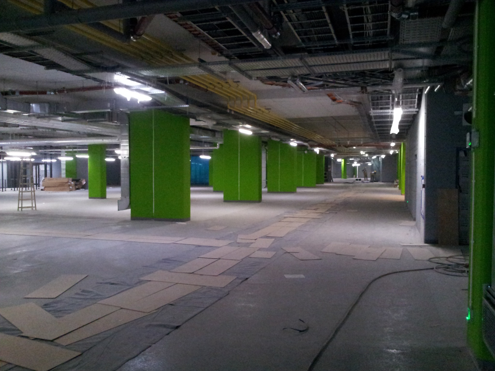 Wnętrze biurowe w trakcie prac wykończeniowych; widoczne zielone kolumny, odsłonięty sufit z instalacjami, podłoga zabezpieczona kartonem i folią.