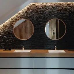 Nowoczesna łazienka na poddaszu z dwoma okrągłymi lustrami i kamienną ścianą z podświetleniem LED. Umywalki wpuszczone w drewniany blat, szare szafki.