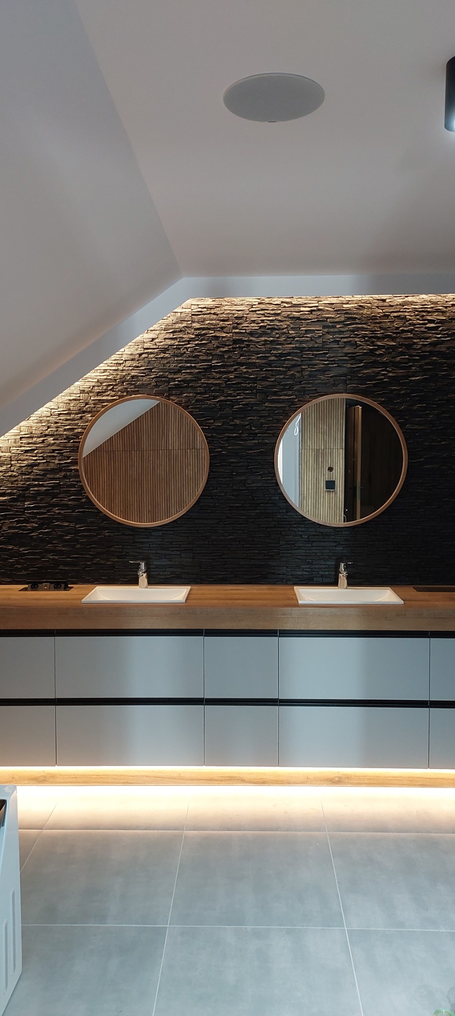 Nowoczesna łazienka na poddaszu z dwoma okrągłymi lustrami i kamienną ścianą z podświetleniem LED. Umywalki wpuszczone w drewniany blat, szare szafki.