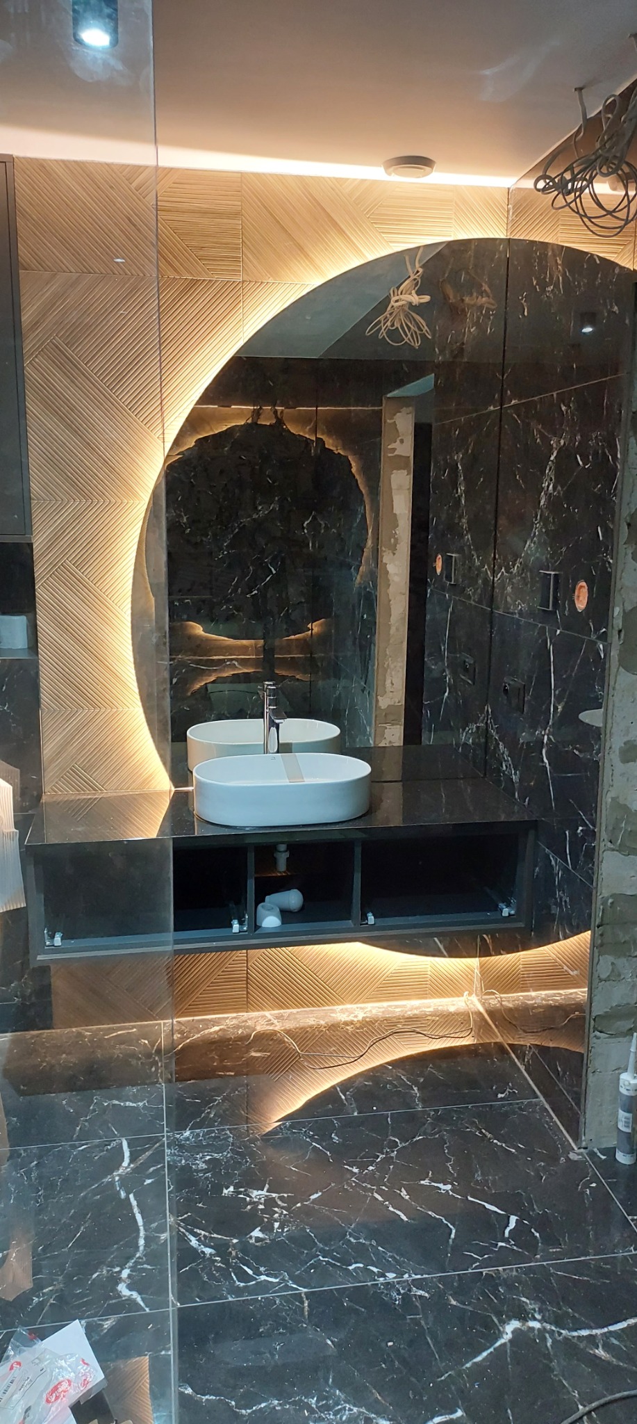 Nowoczesna łazienka w trakcie wykończenia: marmurowe płytki, podświetlane lustro owalne i drewniane panele. Widoczne elementy instalacji i narzędzia.