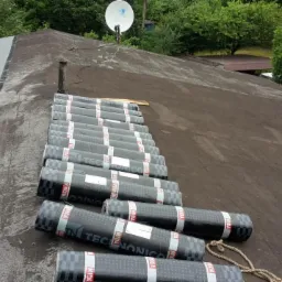 Fragment dachu z częściowo położoną papą termozgrzewalną i przygotowanymi rolkami papy marki Mida, widoczny komin i antena satelitarna.