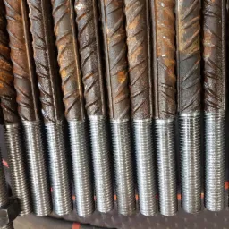 Rząd stalowych prętów z gwintowanymi końcówkami, część z nich pokryta rdzą, ułożonych równolegle na ciemnym tle. Widoczna nakrętka na jednym z prętów.