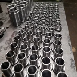 Szereg metalowych tulei z kołnierzem, ułożonych rzędami na szarej, roboczej powierzchni. Widoczne ślady obróbki i drobne niedoskonałości powierzchni.