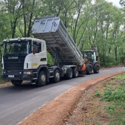 Ciężarówka Scania z podniesioną skrzynią wyładowuje kruszywo na świeżo położony asfalt w Koluszkach, obok pracuje koparko-ładowarka, w tle widoczny las.