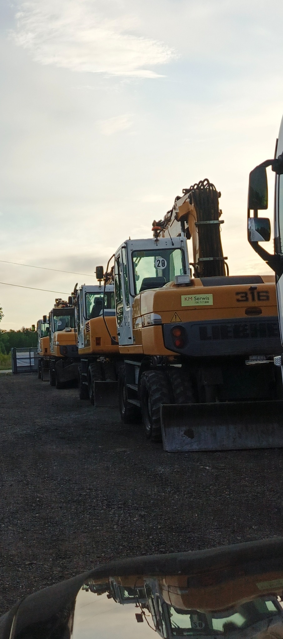 Żółto-białe koparki Liebherr 316 ustawione w rzędzie na żwirowym placu, z widocznym logo KM Serwis. Sprzęt budowlany gotowy do pracy.