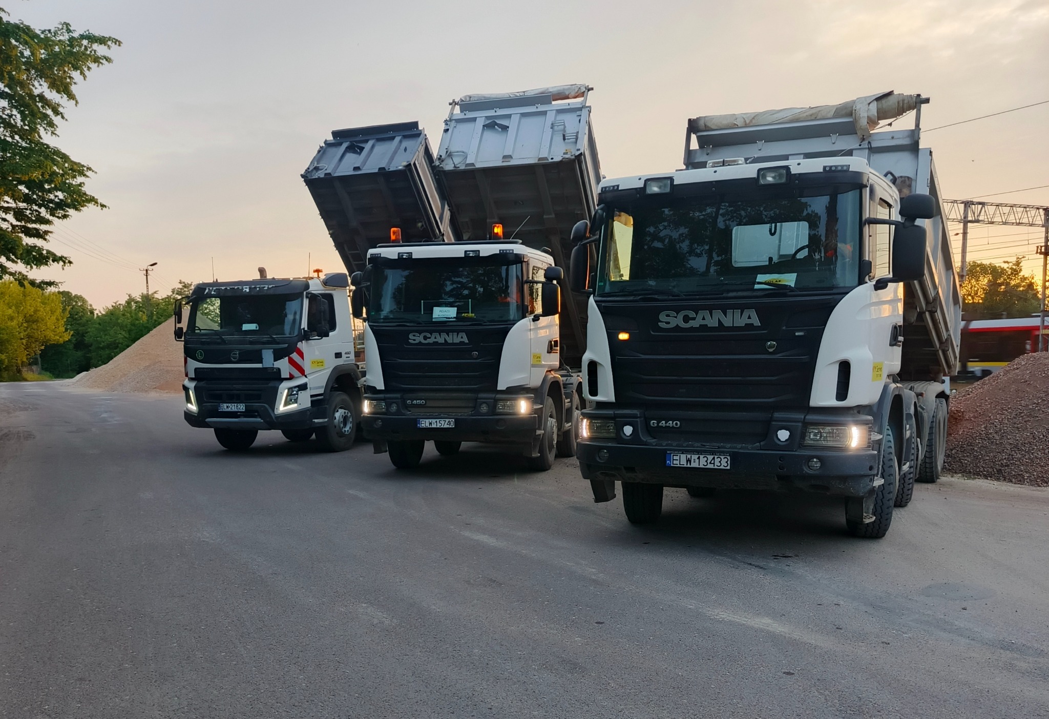 Trzy wywrotki (Volvo i Scania) z podniesionymi skrzyniami na asfalcie, w tle hałda kruszywa i budynek. Dwie Scanie G440 z przodu, Volvo z tyłu. Ujęcie z poziomu jezdni.