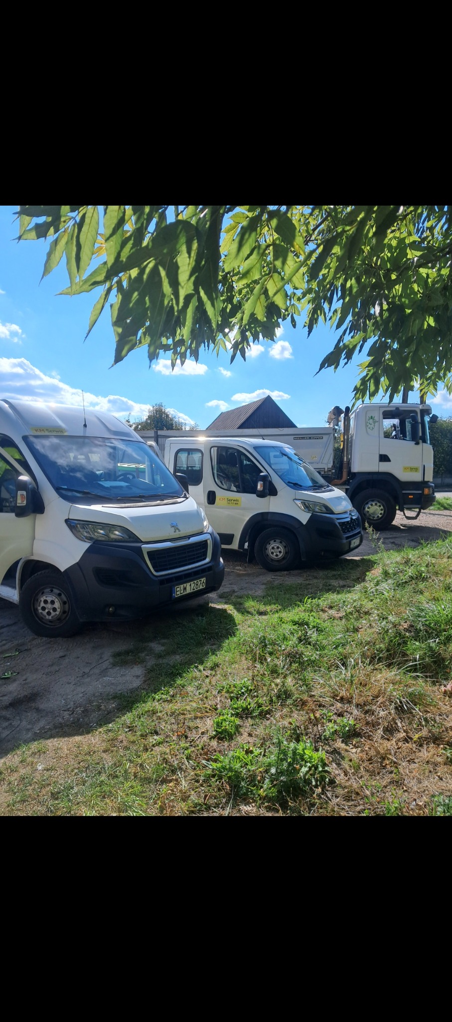 Trzy pojazdy firmy KM Serwis: biały bus Peugeot z rejestracją ELW 12876, bus dostawczy i ciężarówka z kontenerem, stojące na trawiastym terenie w słoneczny dzień.