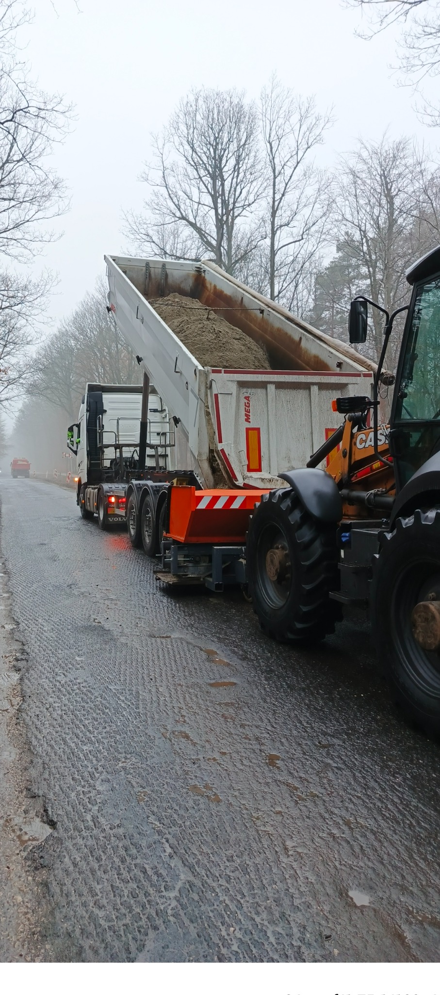 Ciężarówka z otwartą skrzynią wyładowuje piasek na przygotowaną drogę. Obok stoi koparka. W tle drzewa i mgła.