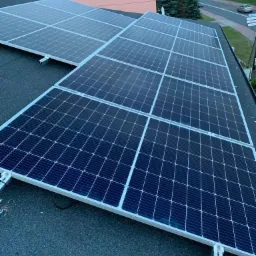 Panele słoneczne zamontowane na dachu domu jednorodzinnego, widoczne metalowe wsporniki montażowe i kable, w tle fragment różowego budynku i ulica.