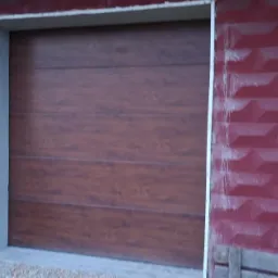 Panelowa brama garażowa w odcieniach brązu, zamontowana w budynku z bordową, teksturowaną elewacją. Widoczne świeże wykończenia wokół ościeżnicy i betonowy zbiornik obok.