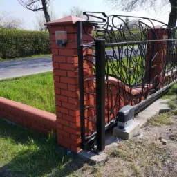 Przesuwna brama wjazdowa z automatyką, czarna, z dekoracyjnym wzorem liści, zamontowana na murku z czerwonej cegły z lampą ostrzegawczą, widoczny napęd bramy na betonowym fundamencie.