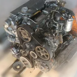 Silnik spalinowy z widocznym paskiem rozrządu, umieszczony na drewnianej palecie w warsztacie.