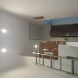 Wnętrze w trakcie remontu: ściany pokryte świeżą warstwą gładzi, widoczne punkty oświetleniowe, drabina aluminiowa, płyta OSB oparta o ścianę oraz fragment sufitu z niebieskim akcentem.