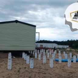 PEP-KOP Roboty Ziemne Usługi Koparko Ładowarką , Minikoparką Wykopy glebogryzarka separacyjna trawniki płyty fundamentowe Różan - Montaż domu modułowego na przygotowanych, betonowych słupach fundamentowych z widocznym logo firmy Pep-Kop w prawym górnym rogu, na piaszczystym terenie pod pochmurnym niebem.