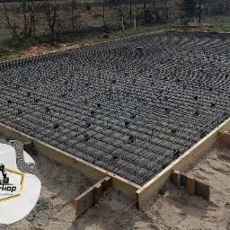 Zbrojenie płyty fundamentowej w drewnianym szalunku, widoczne podkładki dystansowe i logo firmy w rogu zdjęcia.