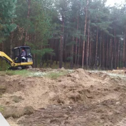 Żółta mini koparka i traktor w trakcie prac ziemnych na zalesionym terenie, widoczne ślady opon i wykopana ziemia, logo firmy w lewym dolnym rogu.